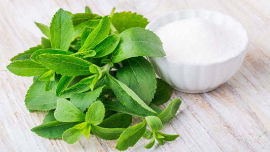 Fresh stevia leaves next to a bowl of stevia powder on a wooden surface.