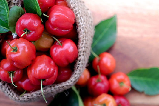 A basket overflowing with fresh Acerola Cherry fruits and green leaves on a wooden surface.
