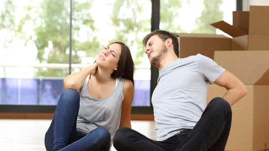 Couple experiencing neck and back discomfort while sitting on the floor in a room with moving boxes.