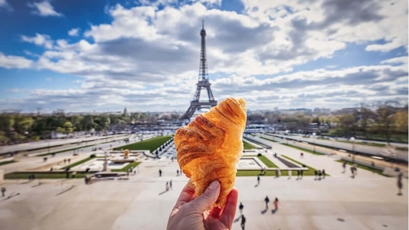 A person holding a croissant in front of the Eiffel Tower symbolizing Health Foods of French People.