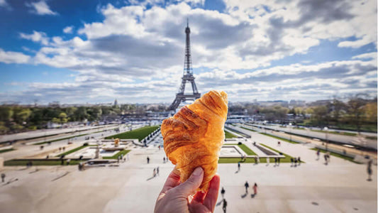 A person holding a croissant in front of the Eiffel Tower symbolizing Health Foods of French People.