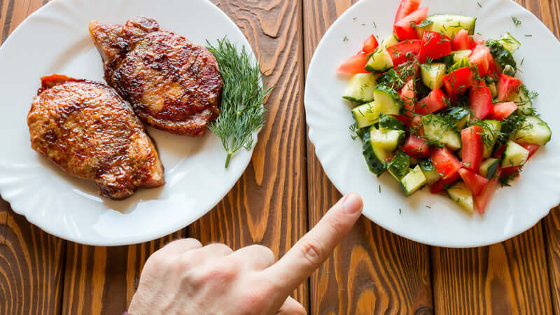 A hand pointing at a plate of salad while meat is on the other plate, illustrating Vegan Against Meat.