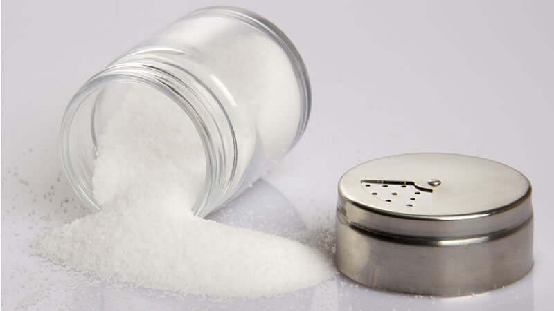 A glass jar spilling white granulated sodium onto a surface next to a metal salt shaker.