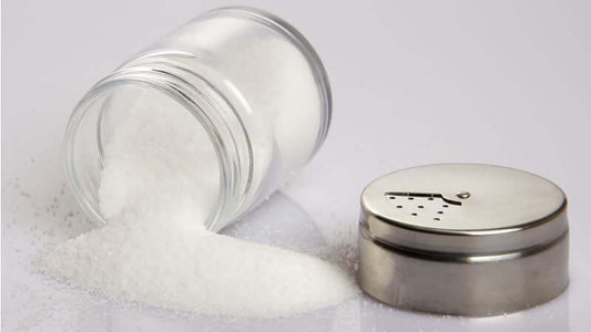 A glass jar spilling white granulated sodium onto a surface next to a metal salt shaker.