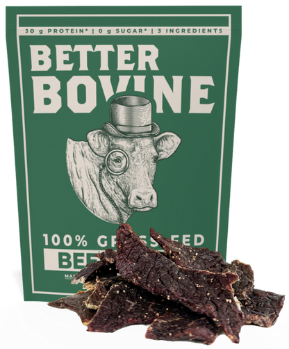 A package of Better Bovine beef jerky stands upright against a plain white background, with several dark brown jerky slices piled in front of the box. The package is dark green with white text and an illustration of a cow wearing a top hat and monocle.