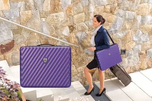 A woman in a business outfit walks up stone steps while carrying a purple quilted briefcase-style bag and a gray rolling suitcase. A large inset close-up at left shows the purple bag front with diagonal quilting and a small circular logo badge.