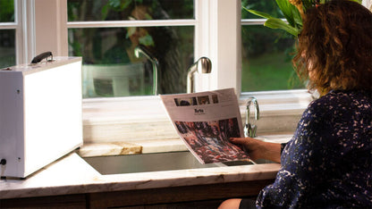 Person reading a magazine by a window with a sink and faucet in the foreground