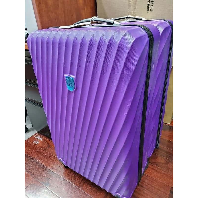 A purple hard-shell suitcase with a textured vertical ribbed design, black zipper, and a small blue shield logo, shown on a wooden floor.