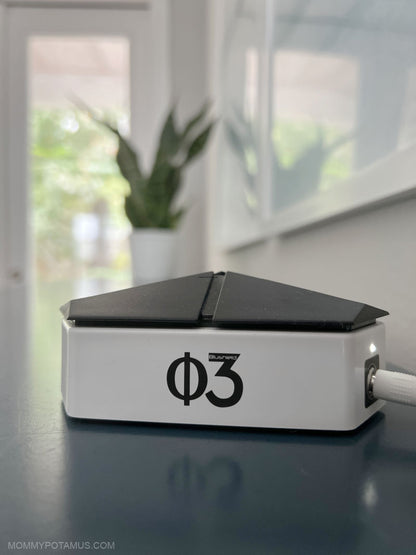 A white rectangular electronic device with a black triangular top and a black phi3 logo on the front, sitting on a dark reflective surface with a blurred indoor background.