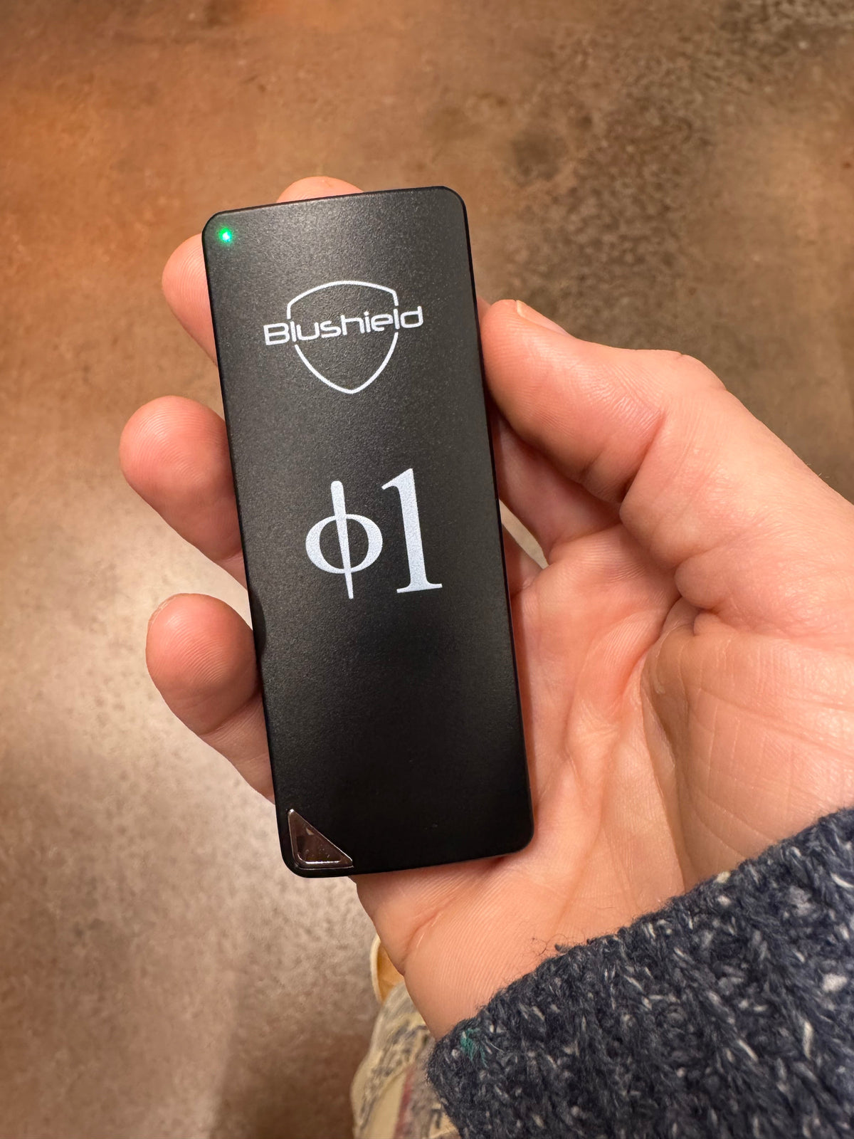 A hand holding a small rectangular black electronic device labeled 'Blushield' and 'phi1' in white text, with a green indicator light on, against a brown textured background.