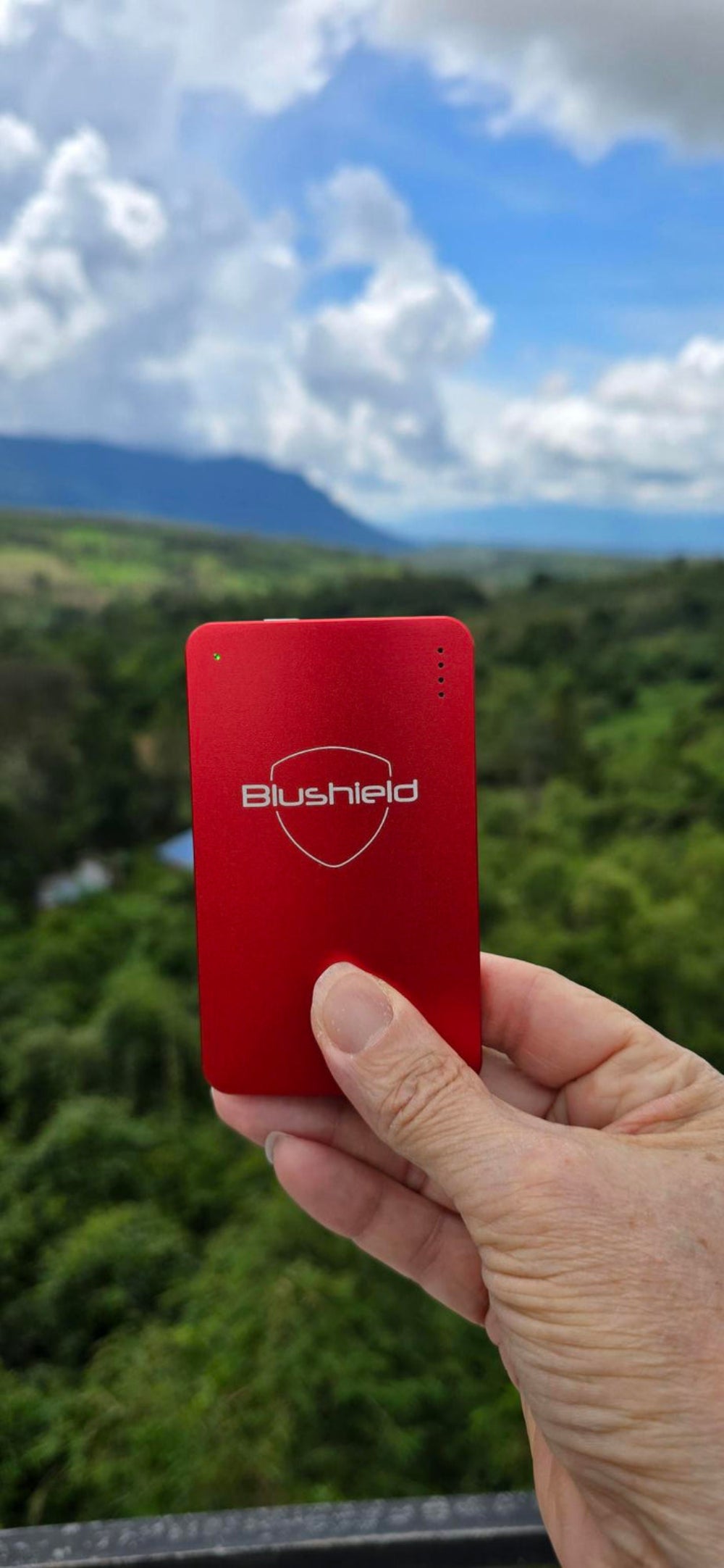 Red Blushield device held in a hand with a scenic background of trees and mountains.