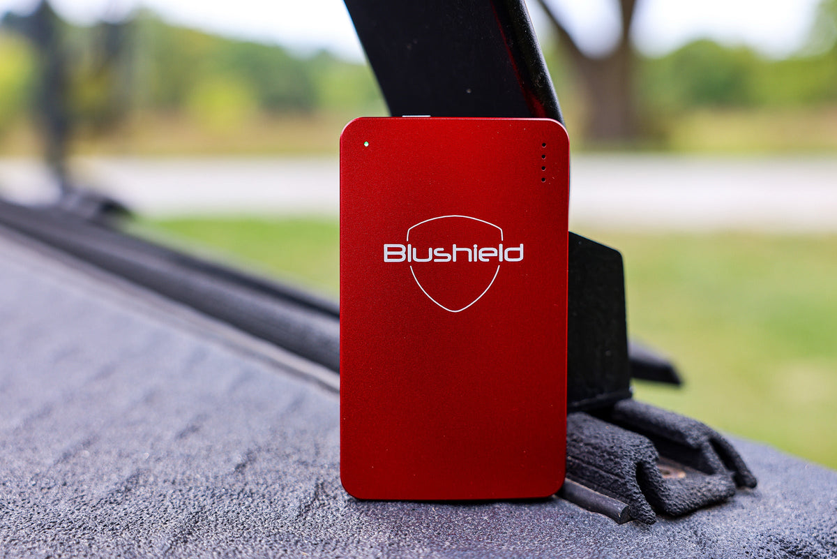 A red rectangular electronic device with the 'Blushield' logo printed in white, mounted on a black strap over a textured gray surface with a blurred outdoor background.