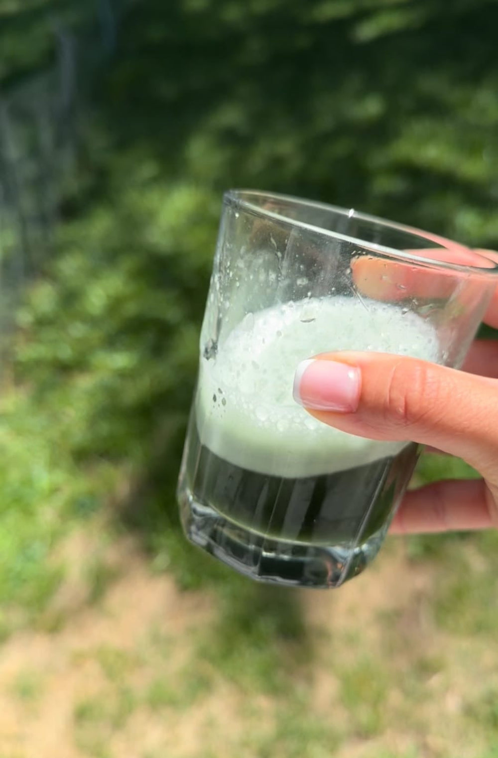 Hand holding a glass with a greenish liquid against a blurred natural background