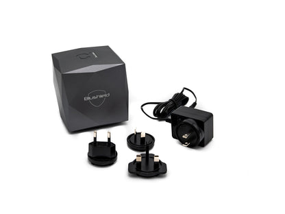 A black rectangular electronic device with a geometric design, shown alongside its packaging box, a power adapter, and three different UK plug types on a white background. The box features the 'Blushield' brand name.