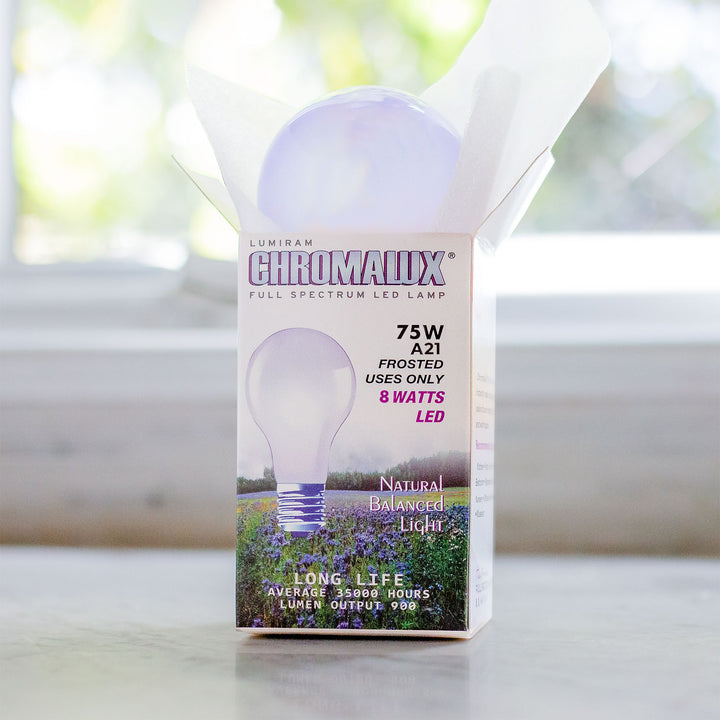 A Lumiram Chromalux 75W A21 full spectrum LED light bulb in its original white packaging box, set on a light-colored surface with a blurred window background.