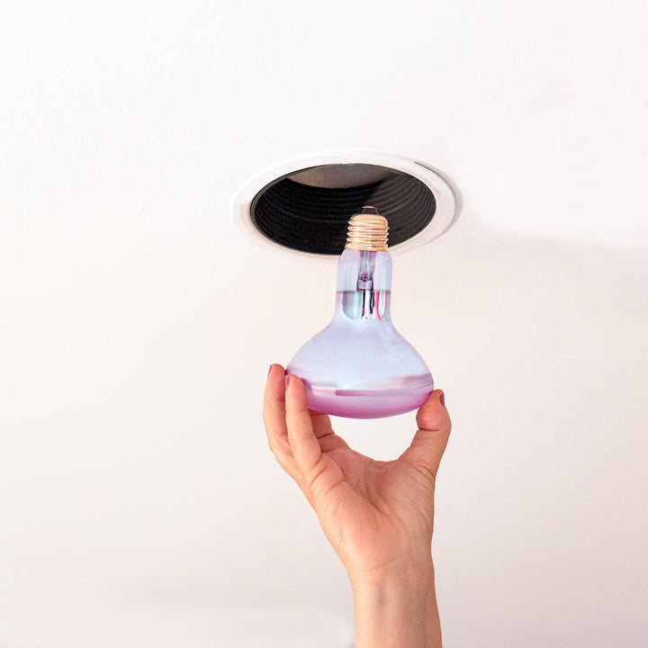 A hand holds a clear glass light bulb with a purple liquid layer and a gold screw base beneath a recessed ceiling light fixture.