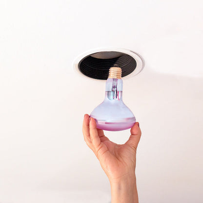 A hand holds a clear glass light bulb with a purple liquid layer and a gold screw base beneath a recessed ceiling light fixture.