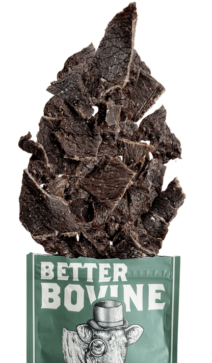 Dark brown beef jerky slices piled on top of a green bag labeled 'BETTER BOVINE' with a cow illustration, isolated on a white background.