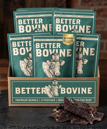 A display box filled with multiple Better Bovine beef tallow pouches, with a few dried beef jerky slices placed in front on a dark tabletop against a rustic brick background.