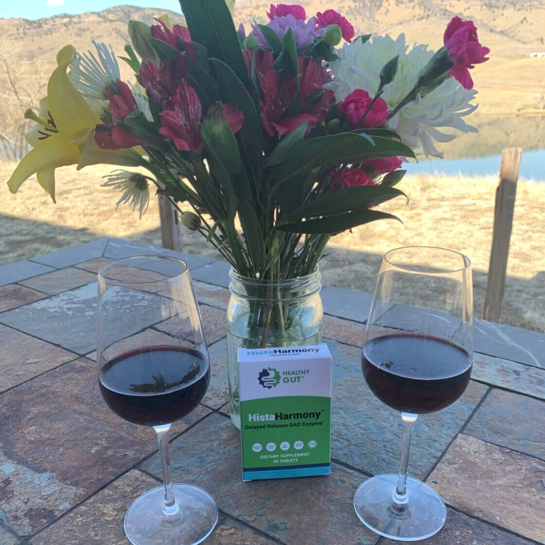Two wine glasses with red wine, a vase of flowers, and a bottle labeled 'Hista Harmony' on a patio table with a scenic background.