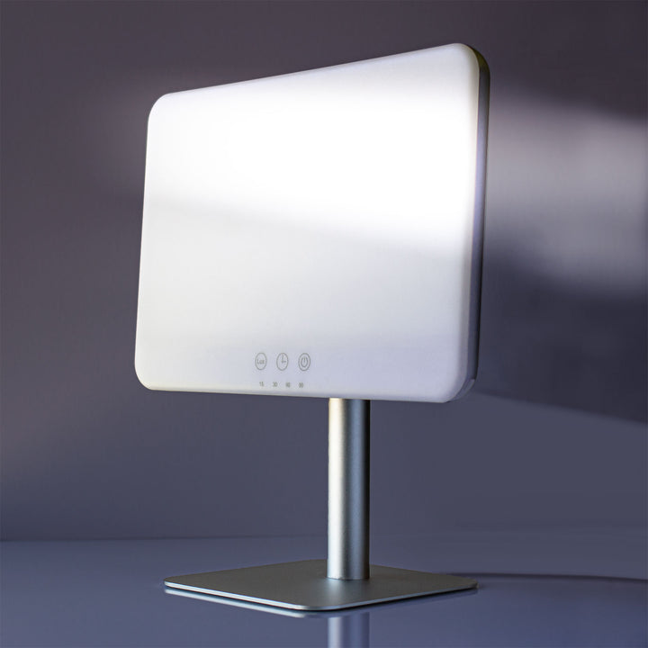 A modern LED vanity mirror with a white illuminated face, mounted on a brushed silver metal stand with a square base, set against a dark gray studio background.