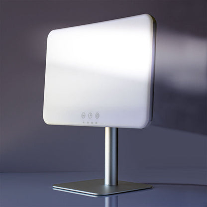 A modern LED vanity mirror with a white illuminated face, mounted on a brushed silver metal stand with a square base, set against a dark gray studio background.