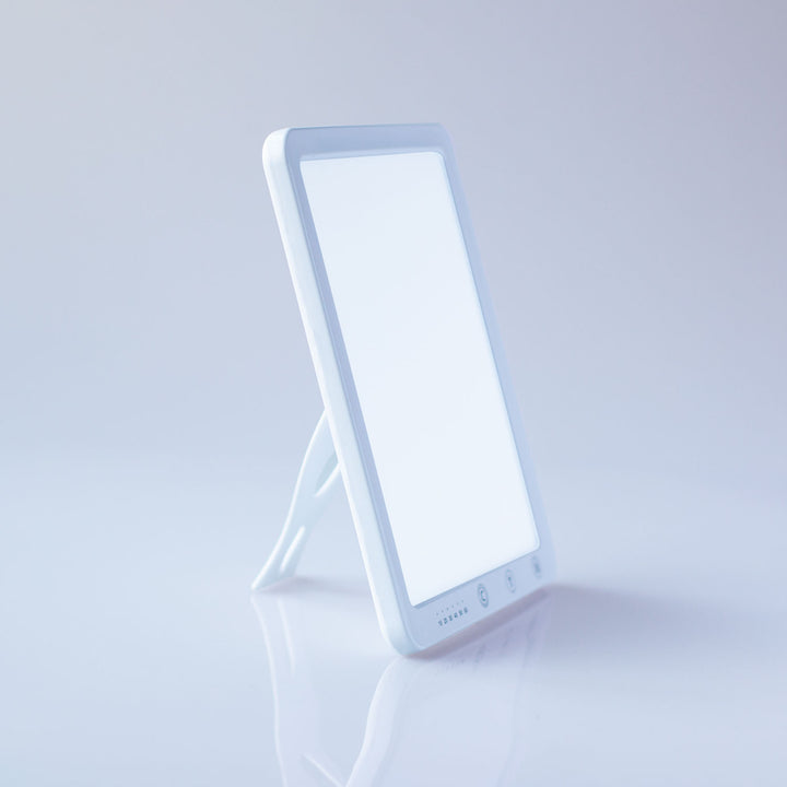 A white LED vanity mirror light with a built-in stand, shown on a clean white reflective surface against a light gray background.