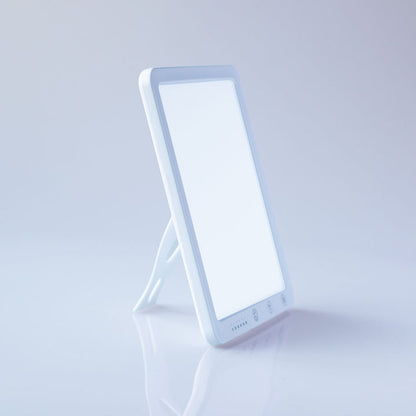 A white LED vanity mirror light with a built-in stand, shown on a clean white reflective surface against a light gray background.