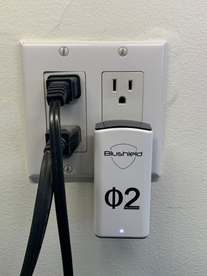 Blushield Φ2 device plugged into a wall outlet with a cable.