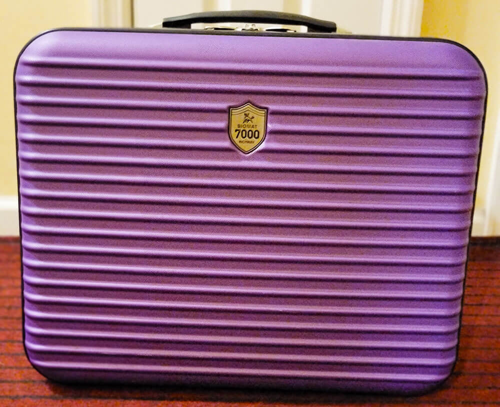 A purple hard-shell suitcase with horizontal ridges and a gold badge on the front, placed on a wooden floor against an indoor background.