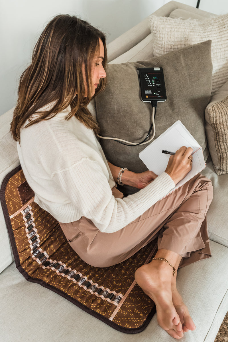 Woman sitting on a patterned mat with a device attached to a pillow, holding a notepad and pen.