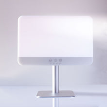 A modern LED vanity mirror with a glowing white illuminated faceplate, a slim silver metal stand, and a square base, shown on a clean white surface against a soft gray background.